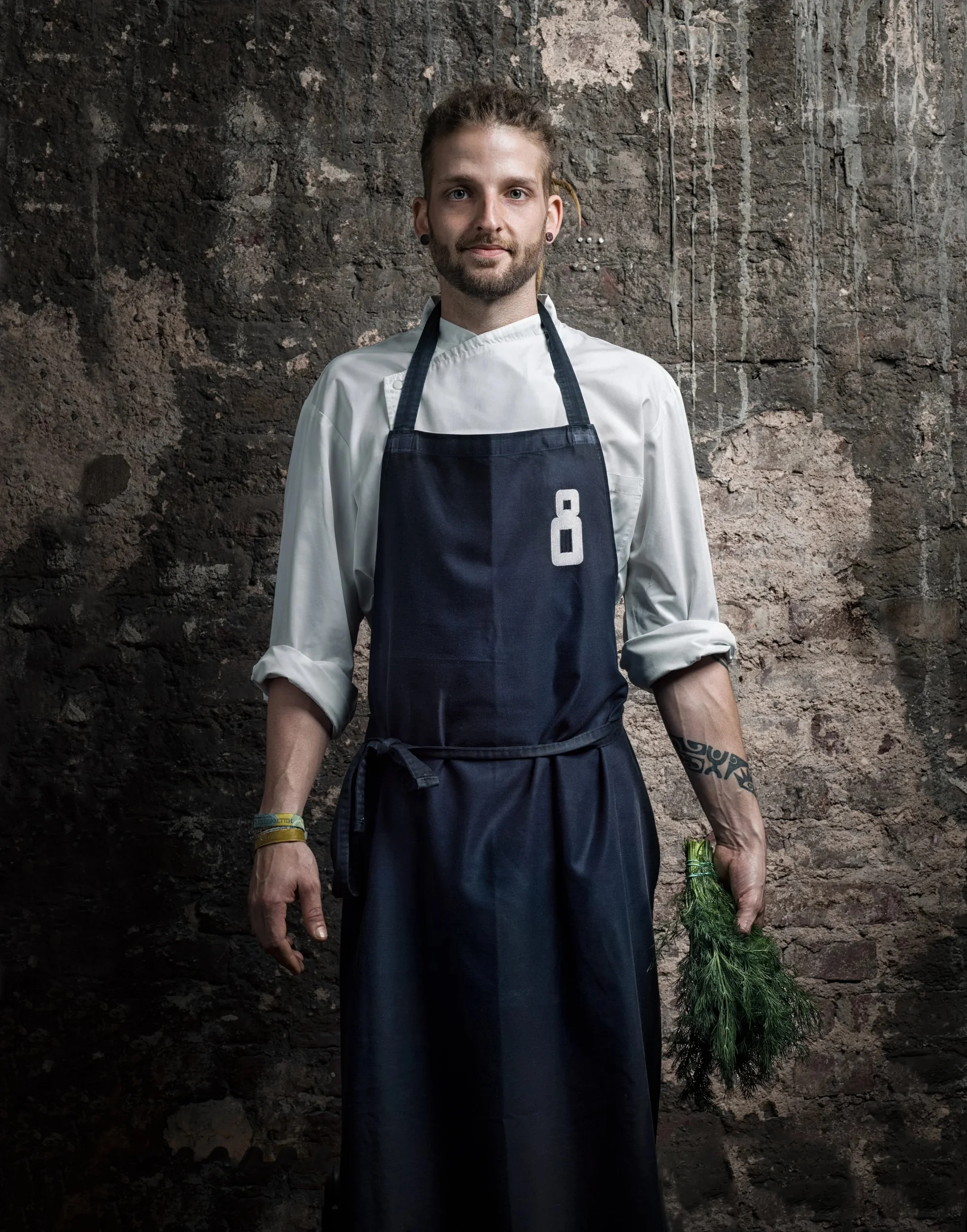 Chef with dreadlocks holding fresh dill against an industrial wall.